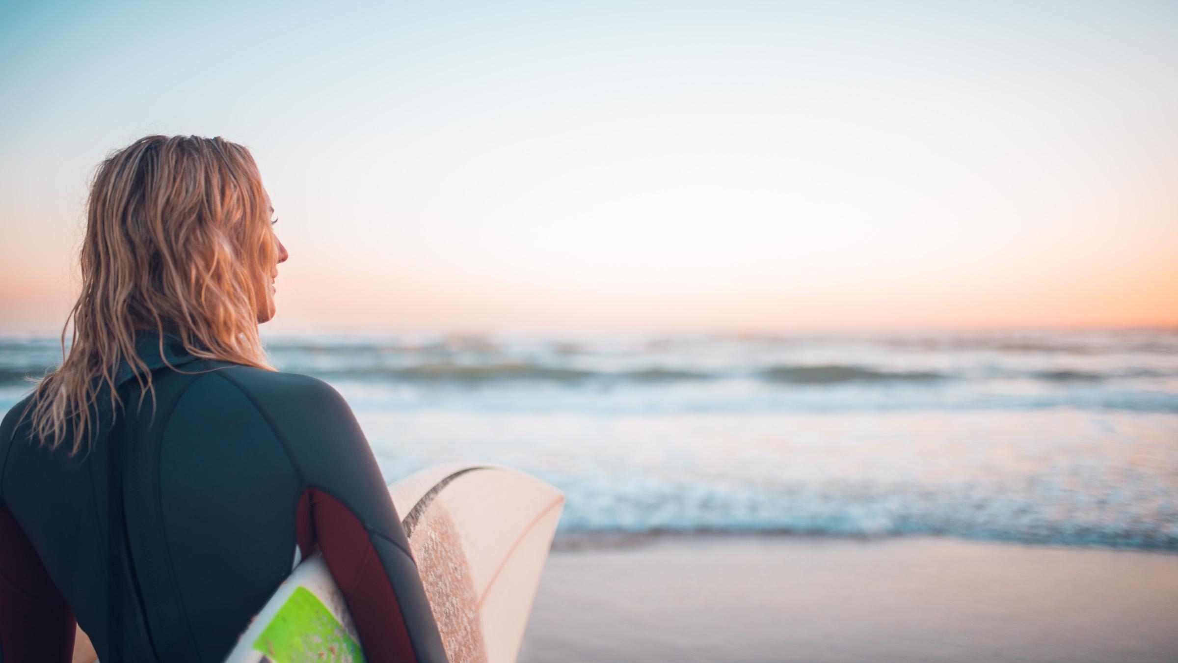 Young surfer standing on the beach at sunrise looking across the waves